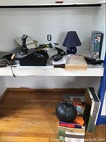 Photo showing multiple items including Logitech joystick, small lamp with blue shade, Pocket Flix handheld players, other miscellaneous items on white shelves and floor