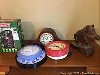 Photo showing mantel clock in wood finish, red alarm clock, three wall clocks with different design borders, Coleman product box, and wooden boar carving.