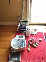 Wide shot showing all items on red carpet including foot spa, shoe stretchers, boxes, curtain rod, and blind.