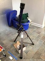 Full view of the blue Tasco Galaxsee telescope mounted on its tripod, with accessories in a box beneath on tiled floor.