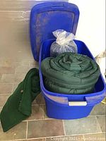 Three green sleeping bags stored with a blue plastic storage tote, one sleeping bag laid out outside the tote. Tote shows dirt marks.