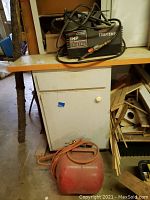 Full view of the Craftsman air compressor on cabinet top with red Airpac compressed air tank and hose on floor below, showing dusty environment.