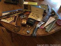 Wide view of lot on wooden table showing two sheathed blades, costume jewelry in boxes, Asian fans, Turtle Ship figurine, silver plate cup, miniature utensils and bowl, and wax decor pieces