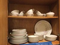 Shelf displaying the cups, creamer, lidded sugar bowl on top shelf, and various plates, saucers and bowls on bottom shelf, all Mikasa My Love china with pink floral pattern.