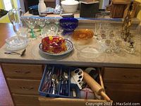 Tabletop displaying glassware collection including beer glasses, crystal bowls in blue and amber colors, a red and clear pressed glass bowl, clear glass platters, a clear glass juicer and pitcher, and a Czech 1970s cut glass decanter with glasses.