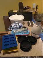 White plastic salad spinner, blue-and-white speckled enamel butter dish, plastic ice trays, black round burger press trays, electric food chopper, metal measuring cup and scoops on countertop