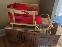 Wooden baby sleigh with red cushion seating installed, yellow pull rope attached, placed on top of original box.