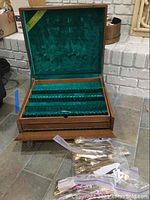Open wooden flatware box with green felt lining, showing empty compartments and multiple brass-toned flatware pieces in plastic bags in front of the box.