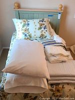 Twin bed set with floral comforter, several folded bedding items on the bed, and a painted twin headboard with pine finials.