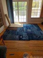 Large blue area rug laid out near sliding glass door with smaller beige rug and rolled carpet remnant visible