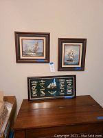 Three framed nautical wall art pieces displayed on a wall and atop a wooden surface showing their overall look and size relations.