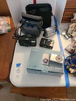 Overview of lot items on table including cameras, camera bags, boxed Canon PowerShot SD400, and accessories.