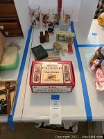 Overview photo of the lot showing grouped vintage items including glasses, coasters, small bottles, playing cards, comb, toothbrush holder, and soap dish arranged on a table with a tag number 24