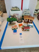 Overview of the collection of 10 miniature house replicas and related collectibles arranged on a table with a white backdrop. Items vary in size and style including cottages, cabins, and a tree figure.