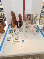 Wide photo of all prayer items grouped together on white table including wooden statues, framed pictures, rosaries, crosses, and figurines.