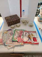 Photo showing miniature hostess set including teapot, sugar bowl, creamer, three cups with saucers, box, and assorted printed handkerchiefs spread flat.