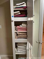 Tall bathroom linen cabinet with multiple shelves holding folded sheets and towels in different colors and sizes.