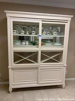 Front view of cabinet showing lighted upper display with mirrored back, floral dinnerware, X-shaped wine rack, and lower panel doors.