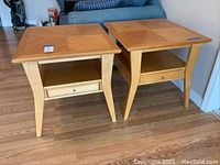 Photo showing two matching light wood end tables with one drawer each and open shelf below, angled legs, geometric patterned top, placed on a wooden floor.