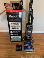 Photo showing Shark Apex vacuum cleaner standing next to its original box, quick start guide, and vacuum bags on wooden floor.