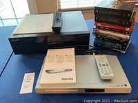 Photo showing Pioneer BDP-51 Blu-ray player stacked over Philips DVP3140 DVD player with remotes and manuals, and a pile of assorted DVDs.