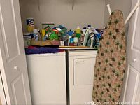 Wide shot showing two laundry appliances topped with numerous cleaning and laundry products and ironing board leaned against wall.