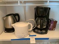 Three appliances on shelf: Hamilton Beach stainless steel kettle, Rival white warmer, Mr. Coffee black coffee maker with glass pot and coffee filters box.