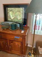 World map framed picture on wooden cabinet with large green candles and brass plates, next to gold tone floor lamp with green shade.