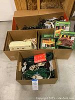 Wide view of three open boxes containing assorted electrical items including extension cords, surge protectors, and boxes of Christmas lights.