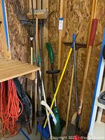 Garden tools leaning against wood wall inside shed including rakes, shovels, trimmer, edger, and broom plus extension cords