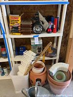 Garden items on white shelving unit including clay pots stacked with white fittings, watering can placed in front, flashlights on upper shelf, box of contractor clean-up bags and pump in original packaging behind pots