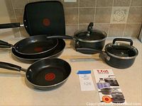 T-fal and Farberware cookware set including fry pans, sauce pans with lids, and griddle arranged on countertop.