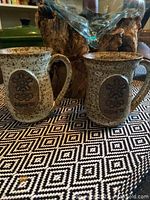 Two beige speckled ceramic mugs displayed on patterned fabric, showing their shape and handles.