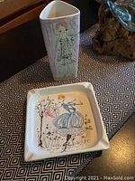 Photo showing both the tall vase with artistic figures and the square decorative dish with painted image of woman dancing outdoors with flowers and tree.