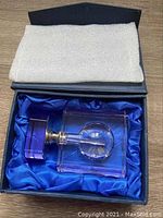 Perfume bottle lying horizontally inside a black box with blue satin lining, showing the square crystal outer structure and purple stopper.