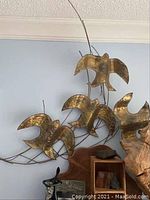 Wide view showing overall sculpture with five metal birds attached to metal rods, hanging on a light blue wall above furniture.