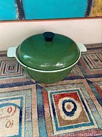 Green enamel pot with lid and black knob shown from an angle on a patterned rug.