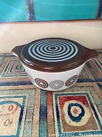 Full view of vintage Pyrex Salton casserole dish with lid showing brown base with blue concentric circles and white glass dish with circular motifs.