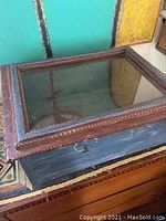 Closed rectangular wooden shadow box with blue painted exterior, decorative carved wooden frame around glass top, and side latch.
