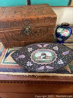 Photo of a carved wood box with detailed carving, metal latch, next to an elongated decorated enamel dish with a central painted building scene and a small cobalt blue vase with pink floral designs on a woven mat.