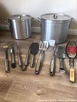 Photo showing two large aluminum stock pots with lids and 8 BBQ utensils including spatula, forks, tongs, basting brush, and fish grill basket all laid out on wooden floor.