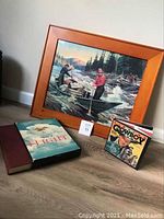 Vintage print titled 'Logging in Mountains' by Philip R. Goodwin, framed in wood, alongside two books on the floor against a wall.