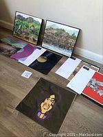 Two framed Hawaiian-themed watercolor paintings on floor against wall with four vinyl items and proofs laid out