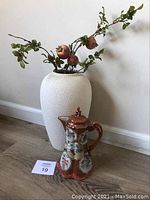 Photo showing white textured decorative vase with pomegranate sprigs and rust gold hand painted Asian tea pot with lid on the floor against a white wall.