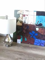 Photo of the table lamp showing crackled glass base, off white shade, and part of abstract floral wall art behind it.