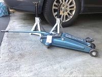 Blue Sears 2 ton hydraulic floor jack shown with handle and wheels, positioned in a garage setting near a vehicle tire. Two white metal wheel stands set behind the jack.