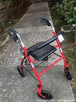 Three-quarter view of red walker with seat and wheels on pavement with plants in background