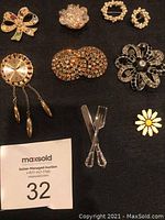 Full view of the 9 pins on a black background, showing various designs including bows, flowers, utensils, and circular shapes with rhinestones and faux pearls.