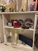 Small white wooden bookcase with two fixed shelves showing chips and scratches in paint; shelves filled with various items not included in sale, including mugs, glass bottles, decorative plates, and masks.