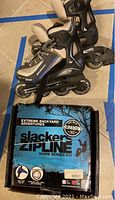 Pair of used inline rollerblades in black and gray with adjustable securing straps resting on the floor next to the boxed Slackers zipline kit.